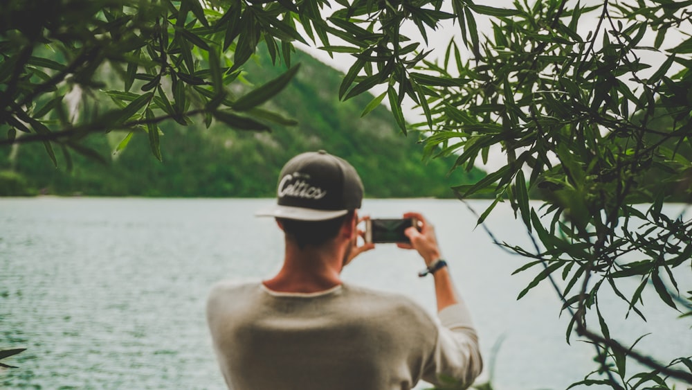 man taking photo of lake using phone near mountain at daytime