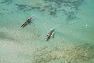 Two long boats with teams of people are paddling through clear turquoise water, seen from an aerial view. There are visible flags in the water, possibly marking a race course.