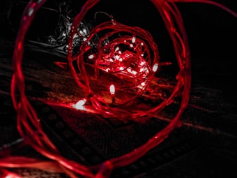 A close-up view of red LED lights intertwined with wires, creating a spiral effect against a dark background. The lights emit a warm, glowing illumination, forming intricate patterns and reflections.