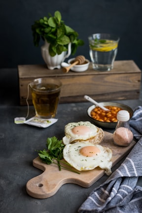 A rustic breakfast setting includes two cooked eggs garnished with herbs on a wooden cutting board, accompanied by fresh parsley. Nearby, a small bowl of baked beans with a spoon, a salt shaker, and a folded striped cloth are visible. In the background, a potted plant, a glass of water with lemon slices, a cup of tea, and a jar of honey complete the scene.