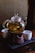 A glass teapot filled with tea leaves is placed on a ceramic warmer, accompanied by two white tea cups on a textured black tray. The setting is dimly lit, creating a cozy ambiance.