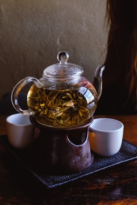 A glass teapot filled with tea leaves is placed on a ceramic warmer, accompanied by two white tea cups on a textured black tray. The setting is dimly lit, creating a cozy ambiance.