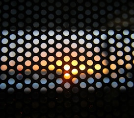 A perforated screen with circular holes is silhouetted against a colorful sunset. The sun is setting on the horizon, casting warm hues of orange, yellow, and hints of blue through the holes.