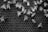 A group of bees is clustered on a honeycomb structure. The hexagonal cells of the honeycomb are visible, and the bees are actively moving about. The image is in black and white, highlighting the contrast between the bees and the darker background of the comb.