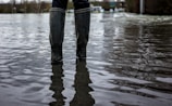 A pair of rubber boots are partially submerged in floodwater, which reflects the overcast sky and surrounding landscape. The scene suggests a flooded area, possibly after heavy rain, with trees and buildings visible in the blurred background.