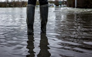 A pair of rubber boots are partially submerged in floodwater, which reflects the overcast sky and surrounding landscape. The scene suggests a flooded area, possibly after heavy rain, with trees and buildings visible in the blurred background.