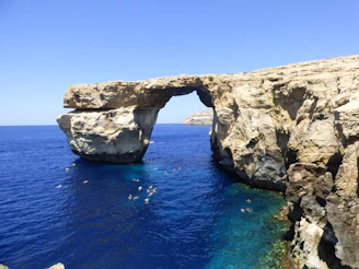 A panoramic shot of the iconic El Arco rock formation with swimmers approaching in the distance.