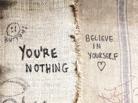 Two contrasting messages are written on a rough, beige burlap fabric. The left side displays the words 'YOU'RE NOTHING' in black ink, along with a sad face and the word 'BUTTFASE' above it. The right side shows a more positive message 'BELIEVE IN YOURSELF' along with a heart symbol, also written in black ink.