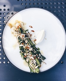 A white plate with a dish of grilled greens topped with sliced almonds and a dollop of cream or soft cheese, set on a perforated dark blue surface.