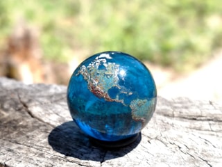 A blue marble resembling a miniature Earth rests on a wooden surface. The globe has landmasses with gold and brown tones, and the background is a blurred natural setting with green and earthy colors.