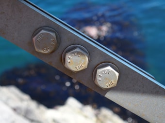 Three metal hexagonal bolts are fastened on a steel beam with an out-of-focus background of blue water and rocks.
