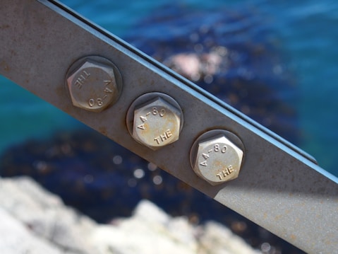 Three metal hexagonal bolts are fastened on a steel beam with an out-of-focus background of blue water and rocks.