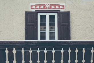 A rustic window with dark wooden shutters on a textured, beige wall. Above the window is an ornamental panel featuring a red geometric pattern. In front of the window, there is a traditional wooden balcony railing with decorative cutouts.
