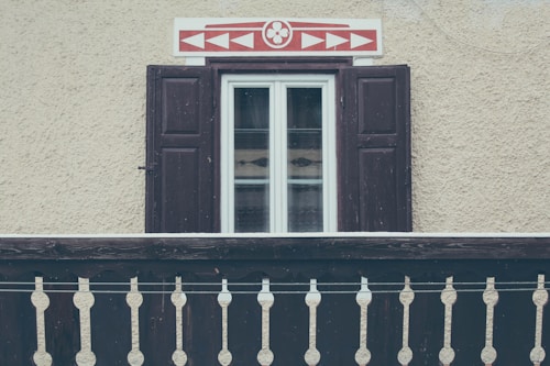 A rustic window with dark wooden shutters on a textured, beige wall. Above the window is an ornamental panel featuring a red geometric pattern. In front of the window, there is a traditional wooden balcony railing with decorative cutouts.