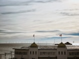 A coastal scene with a large concrete building positioned in the foreground, featuring two domed towers each topped by a flag. In the background, a vast expanse of the ocean is visible, populated by numerous offshore wind turbines under a cloudy sky.