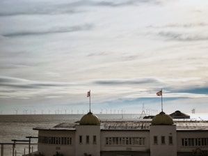 A coastal scene with a large concrete building positioned in the foreground, featuring two domed towers each topped by a flag. In the background, a vast expanse of the ocean is visible, populated by numerous offshore wind turbines under a cloudy sky.