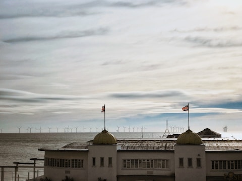 A coastal scene with a large concrete building positioned in the foreground, featuring two domed towers each topped by a flag. In the background, a vast expanse of the ocean is visible, populated by numerous offshore wind turbines under a cloudy sky.