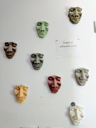 A series of colored sculpted masks are mounted on a white wall. Each mask has distinct facial features and varies in color, including green, brown, yellow, and red. To the right of the masks, a paper is pinned to the wall with the phrase 'People of different colour' written on it. The overall arrangement and theme suggest a commentary on diversity.