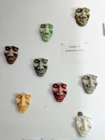 A series of colored sculpted masks are mounted on a white wall. Each mask has distinct facial features and varies in color, including green, brown, yellow, and red. To the right of the masks, a paper is pinned to the wall with the phrase 'People of different colour' written on it. The overall arrangement and theme suggest a commentary on diversity.