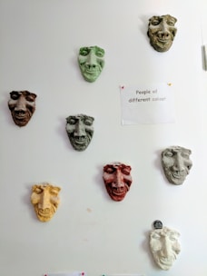 A series of colored sculpted masks are mounted on a white wall. Each mask has distinct facial features and varies in color, including green, brown, yellow, and red. To the right of the masks, a paper is pinned to the wall with the phrase 'People of different colour' written on it. The overall arrangement and theme suggest a commentary on diversity.