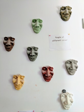 A series of colored sculpted masks are mounted on a white wall. Each mask has distinct facial features and varies in color, including green, brown, yellow, and red. To the right of the masks, a paper is pinned to the wall with the phrase 'People of different colour' written on it. The overall arrangement and theme suggest a commentary on diversity.