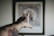 An outstretched tattooed arm reaching towards a blurred framed picture on the wall, with a focus on the intricate tattoo designs on the forearm and hand.