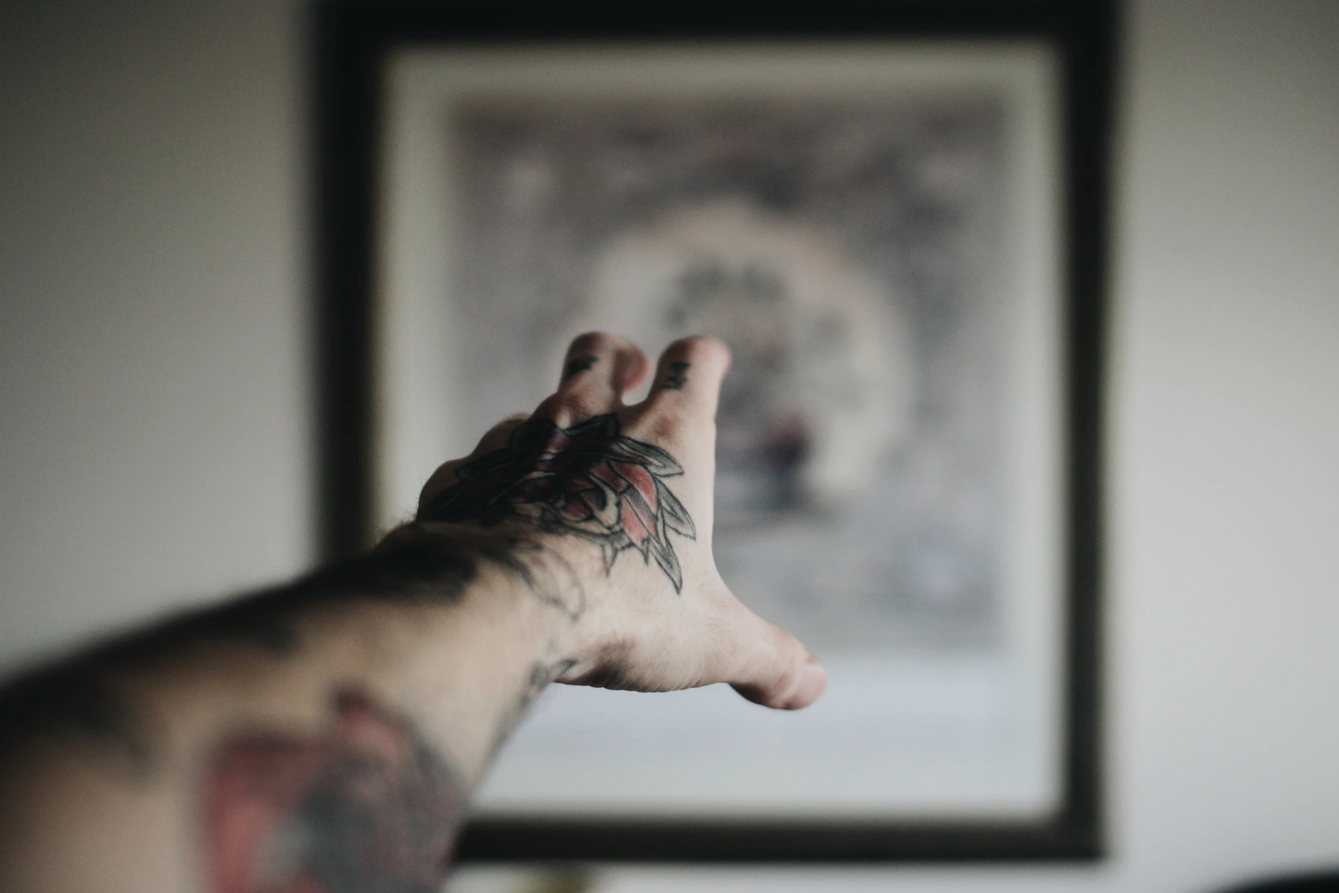 An outstretched tattooed arm reaching towards a blurred framed picture on the wall, with a focus on the intricate tattoo designs on the forearm and hand.