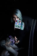 A person with light-colored hair holds a tarot card labeled 'Judgement' in a dimly lit setting. The person's arm and hand are heavily tattooed, and they are seated in a dark, studded chair.
