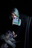A person with light-colored hair holds a tarot card labeled 'Judgement' in a dimly lit setting. The person's arm and hand are heavily tattooed, and they are seated in a dark, studded chair.