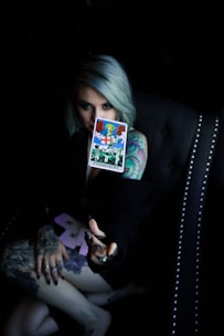 A person with light-colored hair holds a tarot card labeled 'Judgement' in a dimly lit setting. The person's arm and hand are heavily tattooed, and they are seated in a dark, studded chair.