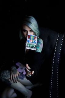 A person with light-colored hair holds a tarot card labeled 'Judgement' in a dimly lit setting. The person's arm and hand are heavily tattooed, and they are seated in a dark, studded chair.