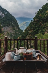 A rustic wooden table set with steaming bowls of traditional Himalayan dishes, framed by a window showing pine trees outside.
