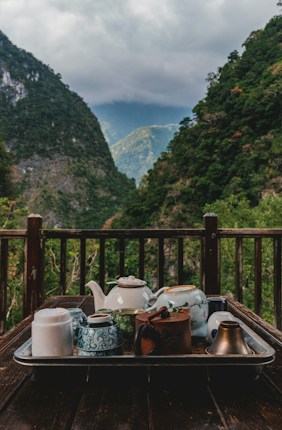 A rustic wooden table set on a deck overlooks a lush, mountainous landscape. Several teapots and cups, each with unique designs, are arranged on a tray. The scene is framed by green hills and a cloudy sky, creating a tranquil atmosphere.