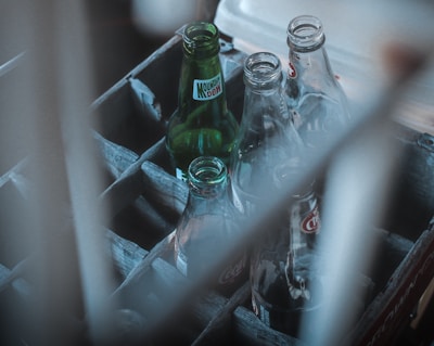 Several empty glass soda bottles, including brands like Mountain Dew and Coke, are placed in a weathered wooden crate. The bottles and crate are partially obscured by an out-of-focus foreground element, giving a rustic and nostalgic feel.