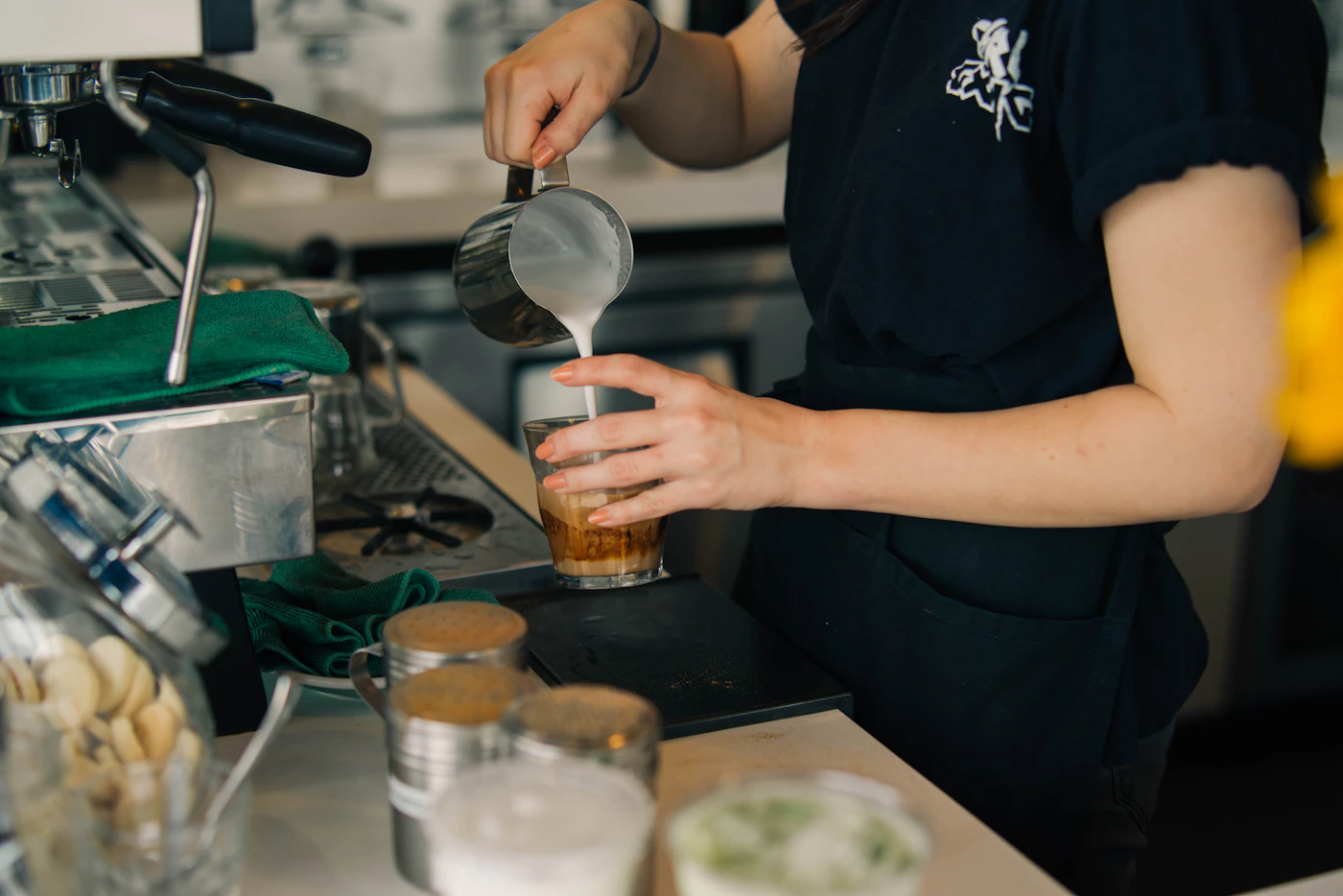 A barista pouring milk into a clear drinking glass
