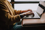 A person typing on a keyboard with LinkedIn open on the monitor in a cozy workspace.