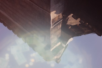 An abstract image featuring a geometric structure with a reflection effect. The structure has a textured surface, possibly resembling stone or concrete, with warm sunlight creating highlights and shadows. Below, a hazy reflection suggests an outdoor environment.