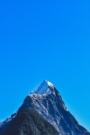 A majestic, snow-capped mountain peak rises steeply against a clear, bright blue sky. The mountain's rugged texture and dark rocky surfaces are visible, with patches of green vegetation near its base.