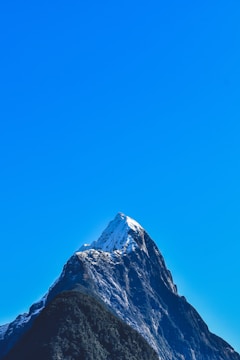 A majestic, snow-capped mountain peak rises steeply against a clear, bright blue sky. The mountain's rugged texture and dark rocky surfaces are visible, with patches of green vegetation near its base.