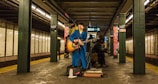 A musician wearing a bright blue suit plays an acoustic guitar in a subway station. Two other individuals are present, one standing behind the performer and another seated nearby, engaged with a phone. The setting is a dimly lit subway platform with tiled walls and visible signage.