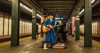 A musician wearing a bright blue suit plays an acoustic guitar in a subway station. Two other individuals are present, one standing behind the performer and another seated nearby, engaged with a phone. The setting is a dimly lit subway platform with tiled walls and visible signage.