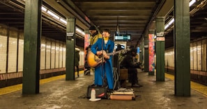 A musician wearing a bright blue suit plays an acoustic guitar in a subway station. Two other individuals are present, one standing behind the performer and another seated nearby, engaged with a phone. The setting is a dimly lit subway platform with tiled walls and visible signage.