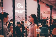 A stylish, indoor social gathering with people interacting in a lively atmosphere. The focus is on two individuals conversing, one holding a wine glass. The background features a Gogo Beauty sign and a wall adorned with photos. The ambiance is cozy and vibrant.
