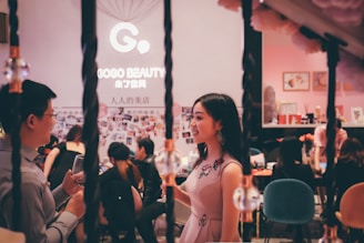A stylish, indoor social gathering with people interacting in a lively atmosphere. The focus is on two individuals conversing, one holding a wine glass. The background features a Gogo Beauty sign and a wall adorned with photos. The ambiance is cozy and vibrant.