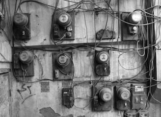 A wall filled with multiple old, dusty electricity meters connected by tangled wires. The meters are mounted in a disorganized fashion on a weathered and cracked concrete surface. Various inscriptions and graffiti are present on the wall, adding to the overall cluttered appearance.