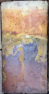 A textured and weathered stone tile with a varied color palette. The surface shows signs of wear and age, featuring irregular patterns and gradients. The colors blend into each other, creating a mosaic-like appearance.