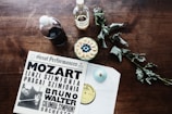 On a wooden table, a vinyl record cover with 'Great Performances' featuring Mozart and Bruno Walter is placed alongside two bottles, one filled with a dark liquid and the other with a clear liquid labeled as sugar. A bowl filled with sliced bananas and blueberries sits nearby, accompanied by a dried leafy plant.