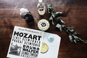On a wooden table, a vinyl record cover with 'Great Performances' featuring Mozart and Bruno Walter is placed alongside two bottles, one filled with a dark liquid and the other with a clear liquid labeled as sugar. A bowl filled with sliced bananas and blueberries sits nearby, accompanied by a dried leafy plant.