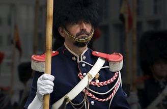 A person dressed in a formal military uniform, featuring a dark blue coat with decorative red accents and gold buttons. The uniform includes a tall fur hat and white gloves, and the person is holding a wooden staff.