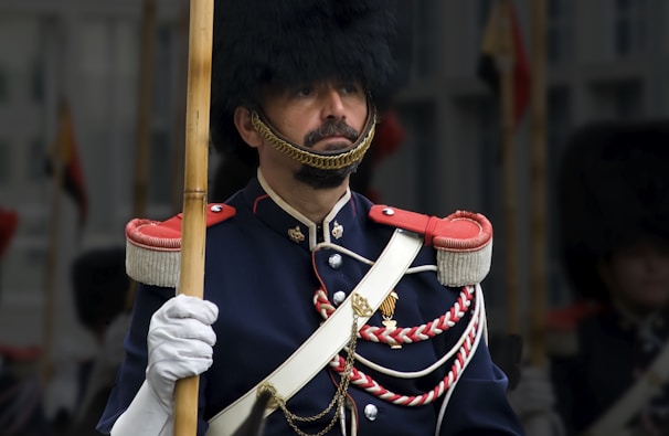 A person dressed in a formal military uniform, featuring a dark blue coat with decorative red accents and gold buttons. The uniform includes a tall fur hat and white gloves, and the person is holding a wooden staff.